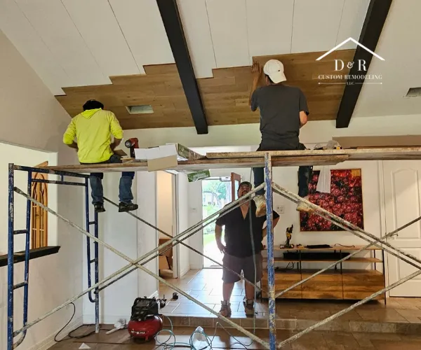 Contractors stand on a high scaffolding to install wood planking on a vaulted ceiling during an interior home renovation.