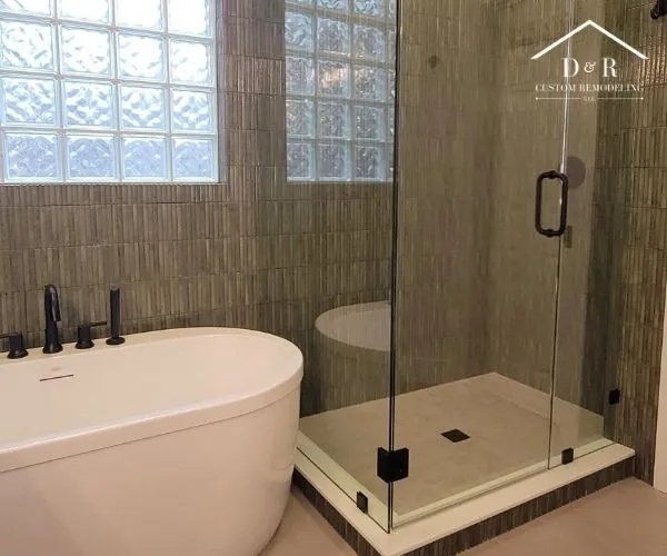 A modern freestanding white bathtub positioned next to a walk-in shower enclosed with glass doors and textured olive-green tile walls.